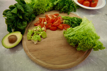 Fototapeta na wymiar High angle view of chopped tomatoes and avocado on a wooden board, variety of greens, lettuce, salad and spinach leaves on a kitchen countertop. Healthy ingredients for raw vegan salad