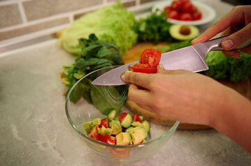High angle view of female hands cutting vegetables with kitchen knife and putting them on glass transparent bowl. Vegan and healthy lifestyle concepts. Preparing raw vegan salad
