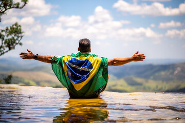 Man with Brazilian flag looking at the horizon. Top of Janela do Ceu Waterfall in Parque Estadual do Ibitipoca (Ibitipoca State Park), Minas Gerais, Brazil - obrazy, fototapety, plakaty