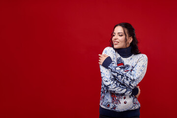 Young woman in a Christmas sweater hugs herself against a red background, smiles and looks to the side. Space for text.