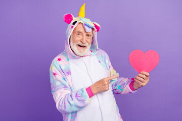 Portrait of attractive cheerful grey-haired man in kigurumi demonstrating heart isolated on violet purple color background