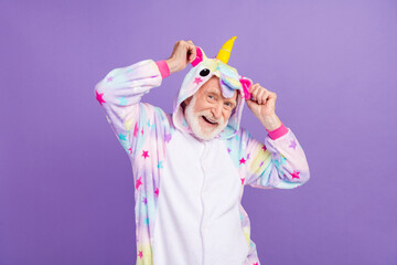 Photo of amazed childish man pensioner unicorn sleepwear dancing arms hands hood smiling isolated violet color background