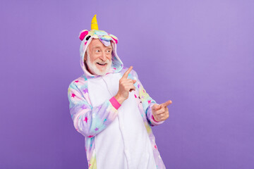 Photo of funny amazed man pensioner unicorn sleepwear looking pointing fingers empty space smiling isolated violet color background