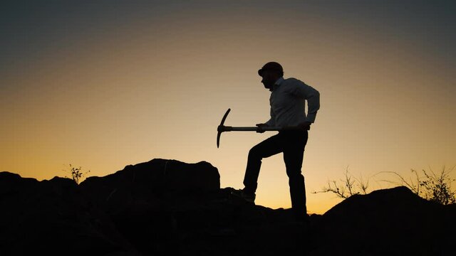 Silhouette man in helmet with pickaxe in his hands stands on rock and hits stone, side view. Man at helmet stands on rock and hits stone with pickaxe