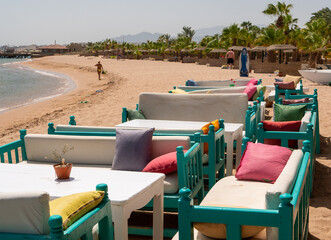 Hurghada, Egypt - September 22, 2021: Beach bar near the red sea. Sofas with multi-colored pillows sit on the sand against a backdrop of thatched umbrellas and green palms.