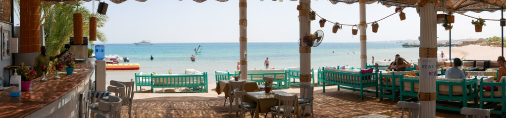 Hurghada, Egypt - September 22, 2021: Panoramic view of the bar on the red sea beach. The bartender pours drinks near the counter. People relax, talk and drink cocktails on sofas at tables.