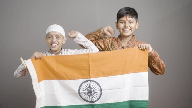 Happy Hindu Muslim kids shouting as India by holding Indian flag on gray background - concept of independence or republic day celebrations, unity in diversity, nationalism and patriotism.