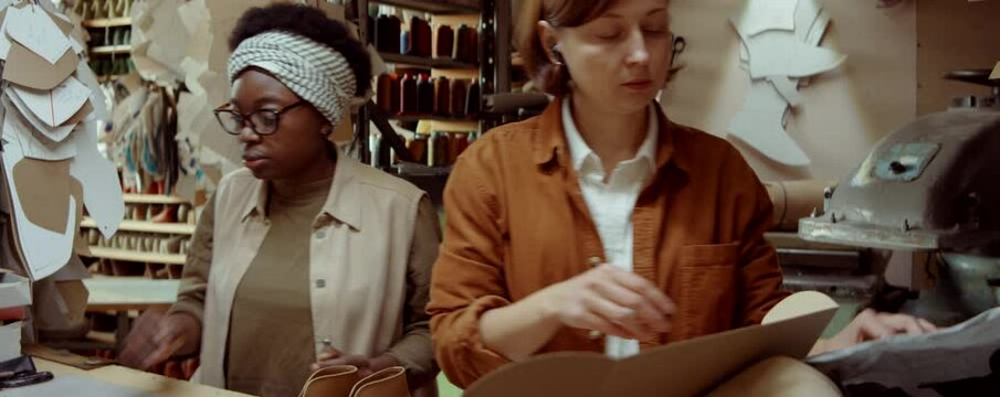 African American woman bringing pair of new shoes and giving them to Caucasian colleague for packing into box while working together in shoemaker workshop