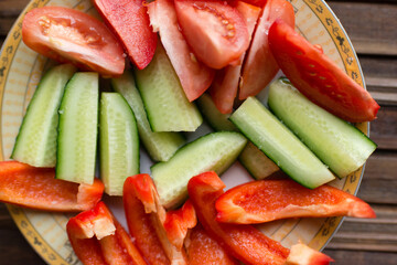 Vegetables on a plate. Tomato, pepper, cucumber cut into pieces. Sprinkled with salt. Vegetables on a plate on a brown wooden background. View from above.