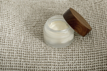 Body cream in glass container with wooden lid on organic linen towel.