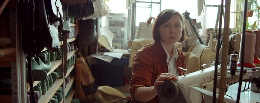 Portrait of woman sitting at desk with sewing machine and posing for camera during workday in shoemaker workshop