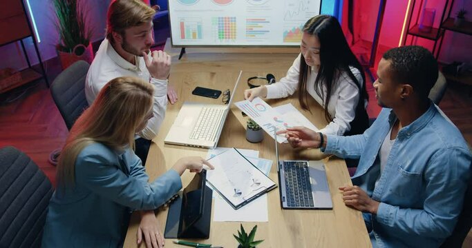 Top view of attractive confident experienced purposeful diverse team of men and women which sitting behind workplace and browsing paper reports and typing on computer in evening office