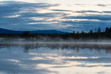 Mist rises from a calm lake at dawn in Kiruna, with mountains in the background near Kebnekaise.