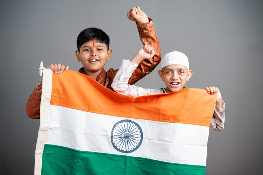 Happy Hindu Muslim kids shouting as India by holding Indian flag on gray background - concept of independence or republic day celebrations, unity in diversity, nationalism and patriotism.