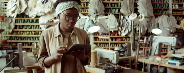 Waist up arc shot of young African American woman working on digital tablet in shoemaker workshop