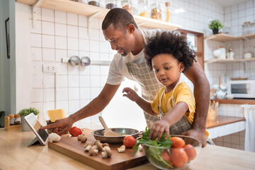 African American Little boy preparing food while his father looking on the digital recipe and using...