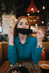 Beautiful young adult woman sits down in a nice restaurant and takes off her protective face mask. She is alone and serious. Coronavirus pandemic and social distancing problems.