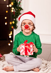 Happy little smiling girl in Santa cap sitting on the blanket in Christmas pajamas with gift box.