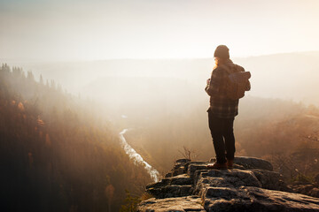 A lone man in a flannel shirt with a backpack on his back stands on a rocky outlook in an autumn landscape during a misty sunset. - obrazy, fototapety, plakaty