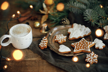 Christmas gingerbread cookies, coffee in stylish white cup, fir branches, warm lights on napkin and rustic wooden table. Atmospheric image. Winter countryside hygge. Happy Holidays!