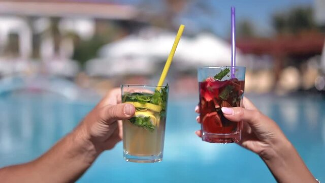 Couple clinking glasses cheers summer cocktails with straw sunbathing swimming pool luxury resort 