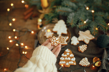 Hand in cozy sweater holding snowflake gingerbread cookie on rustic moody background with fir branches,warm lights, ornaments on table. Winter time. Happy Holidays and Seasons greeting