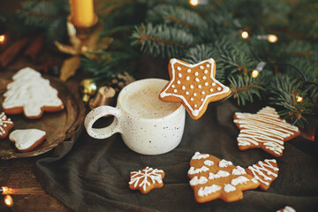 Christmas gingerbread cookies and coffee in stylish white cup, fir branches on napkin and rustic table. Christmas star gingerbread cookie on coffee. Moody atmospheric image. Winter time