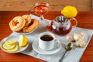 A teapot and a cup and saucer of strong aromatic tea on the table on a napkin next to ginger and lemon jam and donuts.