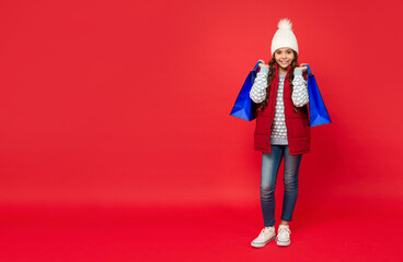 smiling kid in winter hat hold shopping bags on red background with copy space, closeout