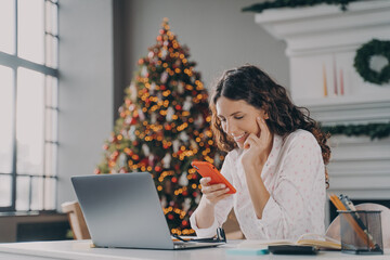 Young happy spanish businesswoman using smartphone while working laptop at home on Christmas