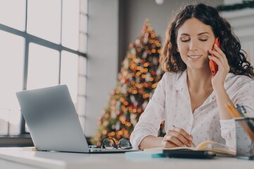 Positive Italian woman with smartphone doing online shopping on laptop during Christmas holidays
