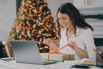 Smiling woman unpacking Christmas gift while sitting at work desk with laptop against xmas tree
