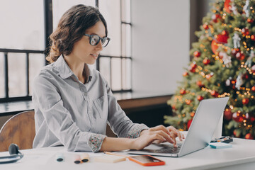 Businesswoman wearing glasses sitting in office near xmas tree and working on laptop