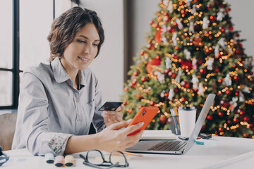 Spanish businesswoman holding credit card, doing online shopping on smartphone during Christmas time