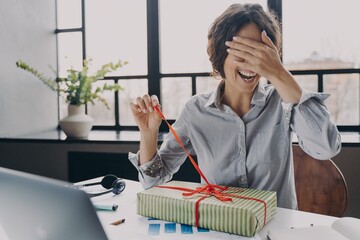 Excited young woman office employee closing eyes with hand while unpacking Xmas present at work