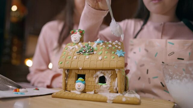 Ginger house with unrecognizable little girl placing sugar icing and woman talking at background. Delicious sweet traditional dessert with Caucasian daughter and mother decorating pastry