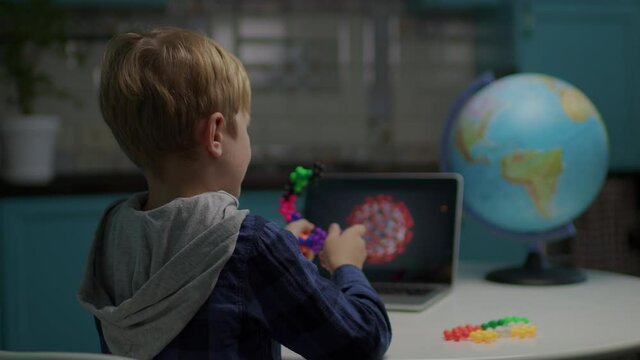Back of school boy playing color puzzle toys making model of virus sitting at home. Kid using tablet computer for modeling virus with toys.