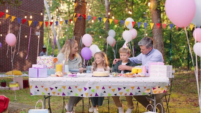Close up of the caucasian little cute girl in a conus celebrating his birthday, preparing to blowing candles on the cake and smiling while sitting with her parents and brother