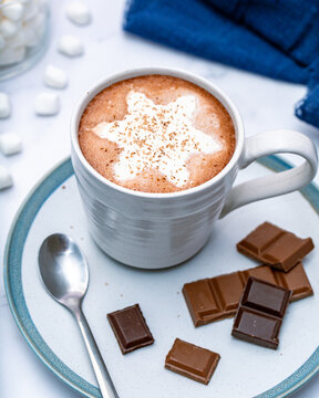 Homemade hot chocolate with whip cream in the shape of a snowflake and chocolate shaving on top in a white mug. Marshmallows are scattered around in the background.