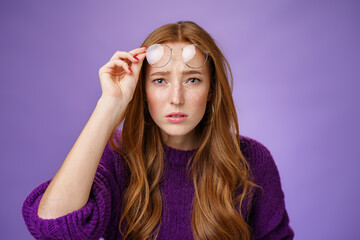 Waist-up shot of intense confused and unsure cute redhead female cannot look without glasses having bad sight taking off eyewear and squinting at camera uncertain, unable to read over purple wall - obrazy, fototapety, plakaty