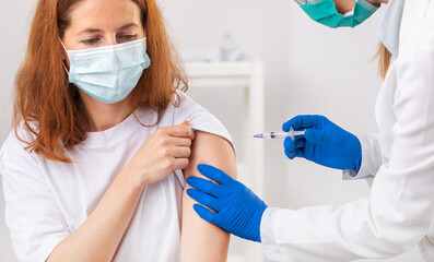 Caucasian woman in face mask sitting at medical cabinet while qualified doctor in gloves doing injection. Vaccination process for coronavirus protection.