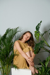 natural looking girl  in a yellow dress sits on a white cube. Tropical plants around, palm, monstera. Minimalism style.
