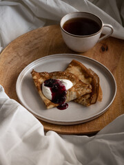 Breakfast in bed, a cup of black tea and thin pancakes with currant jam and sour cream on a beige ceramic plate on a wooden tray on a soft white bed in the morning light