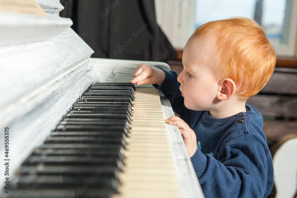 Wall mural Cute one year old redhead baby boy playing white piano keys pressing keys. Adorable male child presses the keys of a white piano