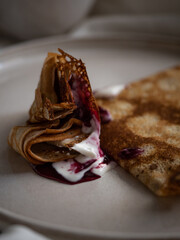 Breakfast in bed, a cup of black tea and thin pancakes with currant jam and sour cream on a beige ceramic plate on a soft white bed in the morning light