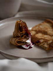 Breakfast in bed, a cup of black tea and thin pancakes with currant jam and sour cream on a beige ceramic plate on a soft white bed in the morning light