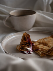 Breakfast in bed, a cup of black tea and thin pancakes with currant jam and sour cream on a beige ceramic plate on a soft white bed in the morning light