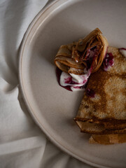 Breakfast in bed, a cup of black tea and thin pancakes with currant jam and sour cream on a beige ceramic plate on a soft white bed in the morning light
