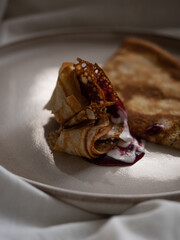 Breakfast in bed, a cup of black tea and thin pancakes with currant jam and sour cream on a beige ceramic plate on a soft white bed in the morning light