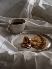 Breakfast in bed, a cup of black tea and thin pancakes with currant jam and sour cream on a beige ceramic plate on a soft white bed in the morning light